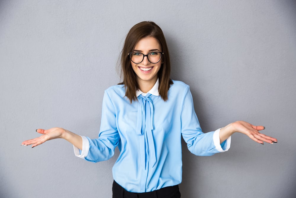 Smiling businesswoman in gesture of asking over gray background. Looking at camera. Wearing in blue shirt and glasses